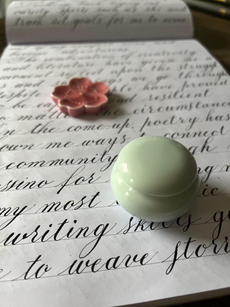 May include: A close-up of a white ceramic container with a lid, sitting on a piece of paper with handwritten text. The text includes the words "community", "poetry", and "writing".