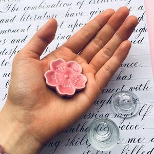 May include: A pink ceramic flower dish with a raised center and a floral design. The dish is sitting in the palm of a hand.
