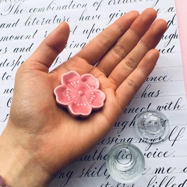 May include: A pink ceramic flower dish with a raised center and a floral design. The dish is sitting in the palm of a hand.