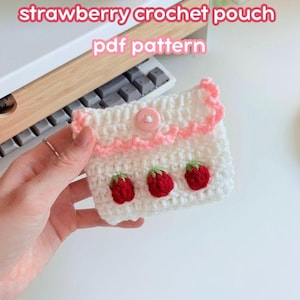 Könnte beinhalten: Eine weiße Häkeltasche mit rosa Besatz und Knopfverschluss. Die Tasche ist mit drei roten Erdbeeren mit grünen Blättern verziert. Der Text "strawberry crochet pouch pdf pattern" ist in Rosa dargestellt.