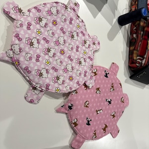 Peut inclure: Deux objets en tissu roses en forme de cochon. L'un présente un motif de personnages Hello Kitty, de nœuds et de fleurs. L'autre a un motif de petits chiens. Les deux ont une bordure rose clair et sont sur une surface blanche.