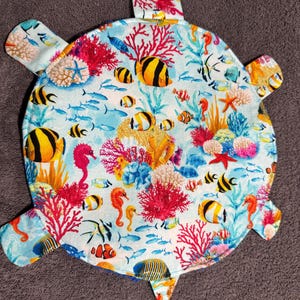 Może przedstawiać: Przedmiot z tkaniny w kształcie żółwia z żywą podwodną sceną. Projekt przedstawia kolorowe koralowce, ryby, koniki morskie i rozgwiazdy na jasnoniebieskim tle. Przedmiot ma cztery przedłużenia w kształcie płetw i kształt głowy.