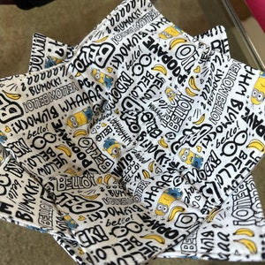 Puede incluir: Un bol de tela blanco con un estampado negro y amarillo con las palabras "bello!" y "popaye" y un personaje de dibujos animados. El bol está doblado y tiene un diseño en forma de estrella.