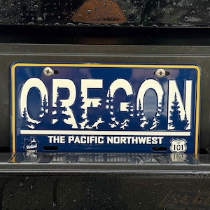 Puede incluir: Placa de matrícula azul de Oregón con letras blancas y un diseño de silueta de bosque. El texto "OREGON" está en la parte superior, con "THE PACIFIC NORTHWEST" debajo. La placa está montada sobre una superficie negra.