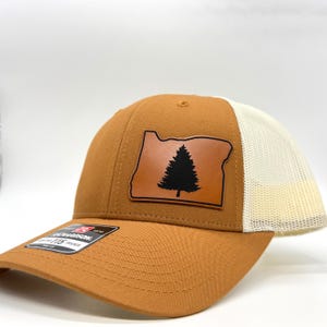 May include: A brown trucker hat with a tan mesh back. The front of the hat features a leather patch in the shape of Oregon, with a black pine tree silhouette. The hat has a curved brim and a tag that reads "Richardson 115 Trucker".