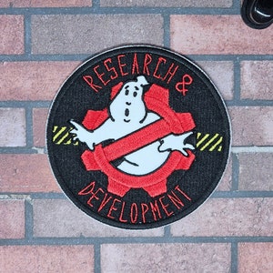 May include: A black and white embroidered patch with a red and white ghost and gear design. The text "RESEARCH & DEVELOPMENT" is written around the edge of the patch.