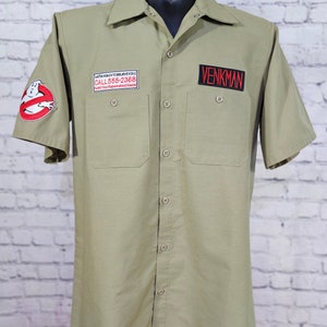 May include: Khaki short-sleeve button-up shirt with two chest pockets. Features a Ghostbusters logo patch on the left sleeve and a "VENKMAN" name patch in red on the right chest. Includes a patch with the text "WE'RE READY TO BELIEVE YOU" and a phone number.