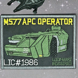 May include: A green and gray embroidered patch with the text "M577 APC OPERATOR" and "LIC #1986" and "LOCK-MART INDUSTRIES". The patch features a green military vehicle with a white number "20" on the side.