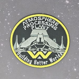 May include: A black and white embroidered patch with a yellow border. The patch features a stylized image of a spaceship with the text "Atmosphere Processing Plant Building Better Worlds".