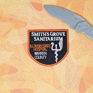 May include: A black and orange patch with the text "SMITH'S GROVE SANITARIUM" and "ILLINOIS STATE HOSPITAL WARREN COUNTY". A gray plastic knife is in the background.