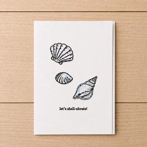 May include: A white greeting card featuring three seashell illustrations in black ink. The card includes the phrase "let's shell-abrate!" in a playful font, perfect for a beach-themed celebration.
