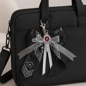 Può includere: Una borsa nera per computer portatile con un accessorio decorativo attaccato al manico. L'accessorio è composto da un fiocco in tessuto nero con motivo paisley bianco, un nastro argentato trasparente e un ciondolo in acrilico a forma di lightstick.
