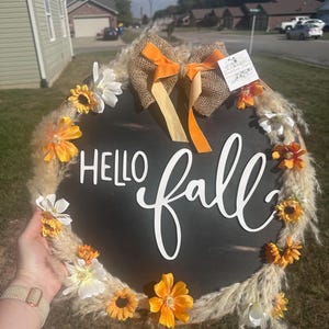 以下が含まれることがあります： 「HELLO fall」の白い文字が入った丸い黒い木の看板。造花のヒマワリ、白い花、乾燥した草で飾られています。オレンジ色のリボンが付いたジュートの弓が上部に飾られています。