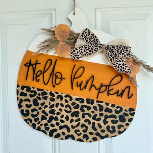 以下が含まれることがあります： レオパード柄、オレンジと白のストライプ、そして「Hello Pumpkin」の文字が書かれた木製のかぼちゃのドアハンガー。ハンガーは、麻の弓と乾燥した葉で飾られています。