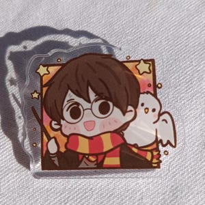 Puede incluir: Un charm de acrílico transparente con una ilustración de dibujos animados de un personaje con cabello castaño, gafas y una bufanda roja y amarilla, sosteniendo una varita. Un búho blanco está posado en el hombro del personaje. El charm tiene forma cuadrada con bordes redondeados y estrellas.