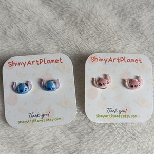 Könnte beinhalten: Zwei Paar Ohrstecker mit einem Cartoon-Charakter-Design. Ein Paar ist blau, das andere rosa. Jedes Paar ist auf einer weißen Karte mit dem Text "ShinyArtPlanet" und "Thank you!" abgebildet.