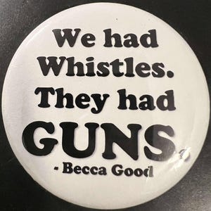 May include: A white button with black text that reads "We had Whistles. They had GUNS." The button also includes the name "- Becca Good." The button is round and has a glossy finish.