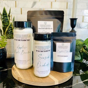 May include: A collection of fabric freshening powders in glass jars and black pouches. The jars are labeled "Apples + Maple Bourbon" and "Seaside Orchids." The pouches are labeled "Egyptian Cotton" and "Cashmere." The products sit on a wooden serving board.