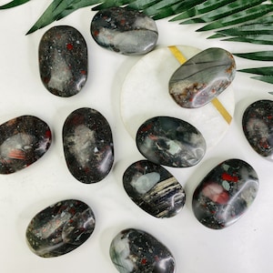 May include: A collection of ten smooth, polished, oval-shaped stones with red, black, and white patterns. The stones are arranged on a white surface with a green leaf in the background.