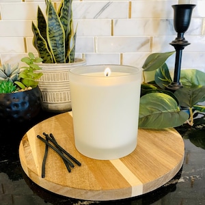 May include: A lit white candle in a frosted glass jar sits on a round wooden serving board with a light wood and white stripe design. Several burnt matches are next to the candle. Decorative plants and a black candlestick holder are in the background.