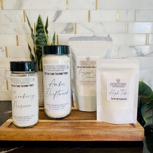 Puede incluir: Cuatro recipientes de polvo ambientador de telas sobre una tabla de madera. Los recipientes incluyen dos frascos de vidrio con tapas negras y dos bolsas blancas. Los frascos están etiquetados como "Cranberry Prosecco" y "Amber Driftwood". Las bolsas están etiquetadas como "Pineapple Coconut" y "High Tide".