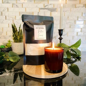 May include: A lit, dark red candle in a clear glass jar sits on a wooden coaster next to a black bag labeled "Complete Candle Making Kit" with the text "Fallen Leaves". A tall, thin candle burns in a black candlestick.
