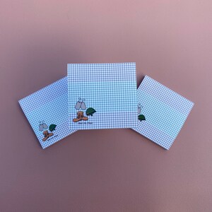 May include: Three square notepads with a blue and white gingham pattern. Each notepad features a cartoon illustration of military boots, a helmet, and dog tags. The notepads are arranged on a solid mauve background.