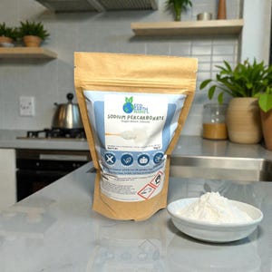 May include: A brown paper bag of Eco Earth Market Sodium Percarbonate, a granular oxygen bleach cleaner. The bag has a blue and white label with product information and icons. A small white bowl contains a pile of the granular powder. The background is a kitchen.