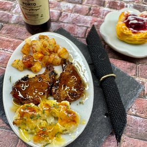 May include: A white plate with two grilled pork chops, roasted potatoes, and cauliflower with a brown sauce. A small white plate with a pastry filled with red jam is in the background.