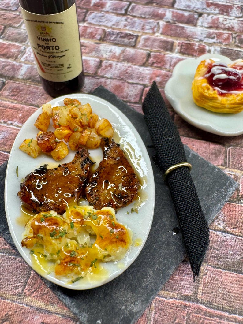 May include: A white plate with two grilled pork chops, roasted potatoes, and cauliflower with a brown sauce. A small white plate with a pastry filled with red jam is in the background.