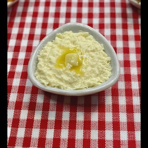Peut inclure: Un petit bol blanc ovale rempli d'une trempette crémeuse jaune pâle, surmontée d'une petite noix de beurre. Le bol est posé sur une nappe à carreaux rouges et blancs, évoquant un cadre décontracté.