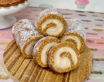 Op de afbeelding: Vier mini pumpkin spice rolletjes met roomkaasvulling, bestoven met poedersuiker, op een houten snijplank.