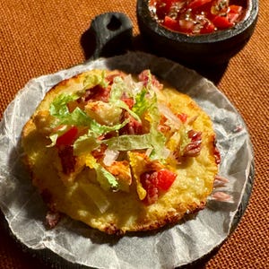 May include: A round, golden-brown flatbread topped with shredded lettuce, diced tomatoes, and what appears to be cooked meat. A small, dark bowl of salsa sits beside the flatbread on a dark, round serving board.