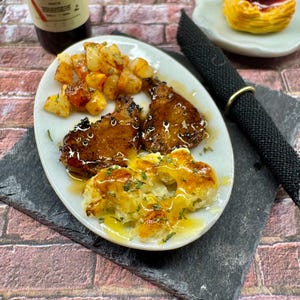May include: A white plate with two pieces of grilled meat, roasted potatoes, and a side of creamy mashed potatoes with a brown sauce. The plate is on a black slate surface. A bottle of wine and a pastry are in the background.