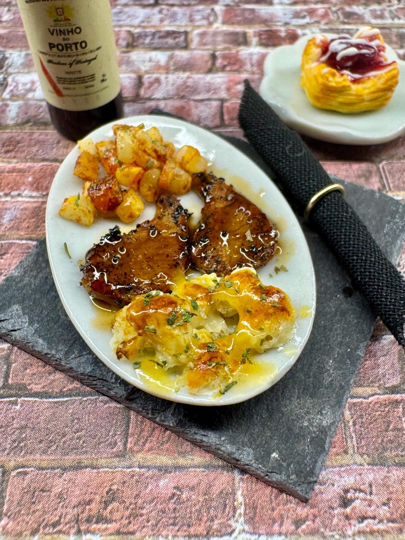 May include: A white plate with two pieces of grilled meat, roasted potatoes, and a side of creamy mashed potatoes with a brown sauce. The plate is on a black slate surface. A bottle of wine and a pastry are in the background.