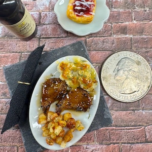 May include: A miniature plate with a meal of two small pieces of meat, mashed potatoes with gravy, and roasted potatoes. A small bottle of wine and a pastry with red jam are also pictured.