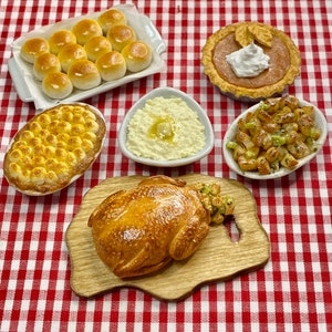 May include: A Thanksgiving dinner table setting with a roasted chicken, mashed potatoes, stuffing, rolls, pumpkin pie, and green bean casserole. The table is covered in a red and white checked tablecloth.