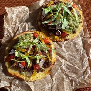 May include: Two open-faced tacos on brown parchment paper. The tacos have a golden-brown base, topped with ground meat, guacamole, chopped tomatoes, onions, and shredded lettuce. The background is a dark brown surface.