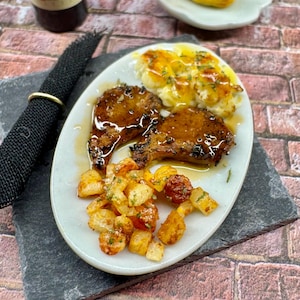 May include: A white plate with two pieces of grilled meat, a side of mashed potatoes with a brown sauce, and a side of roasted potatoes. The meat is glazed with a brown sauce and the potatoes are seasoned with herbs.