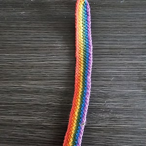 May include: A rainbow friendship bracelet made with colourful threads. The bracelet is tied in a knot at the top and has a long tail.