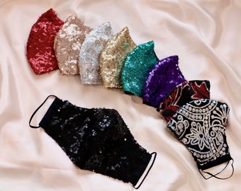 May include: A collection of sequined face masks in various colors, including red, silver, gold, teal, purple, and black. The masks are arranged on a satin surface, showcasing their shimmering texture and design. Each mask has black ear loops.