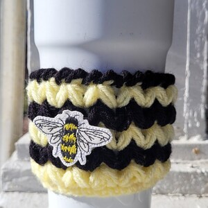 May include: A white tumbler with a black and yellow crocheted cozy featuring a bee applique. The cozy is made of alternating stripes of black and yellow yarn. The text "Made by Mickie Geesaman" is visible.