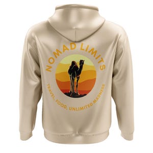 Könnte beinhalten: Beigefarbener Hoodie mit einem runden Grafikdruck eines Kamel-Silhouetten in einer Wüstenlandschaft. Die Grafik lautet "Nomad Limits" mit dem Text "Travel, Food, Unlimited Madness", der den Kreis umgibt.