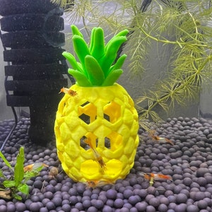 May include: A yellow pineapple-shaped aquarium decoration with a green top and a honeycomb pattern. The decoration is surrounded by small, dark brown gravel.