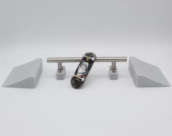 May include: A miniature skateboard with a graphic of an astronaut, leaning against a silver metal rail. Two gray foam ramps flank the rail. The skateboard is black with white wheels. The set is designed for fingerboarding.