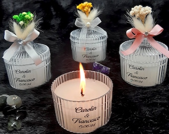 May include: Three clear glass candle jars with white lids and a white ribbon tied around the lid. Each jar has a small bouquet of dried flowers on top. The jars are labeled with the text "Carolin & Francesco 5.06.24". A lit candle is in front of the jars.