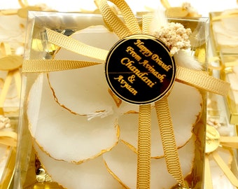 May include: A square clear gift box containing white, round, flat items with gold-painted edges. The box is tied with a gold ribbon and features a round black tag with gold text reading Happy Diwali From Avinash, Chandani & Avyaan.