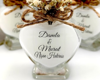 May include: A heart-shaped glass bottle filled with white wax, featuring a jute twine bow tied around the neck and a bouquet of dried white baby's breath flowers. The glass is printed with the black text Damla & Murat Nişan Hatırası.