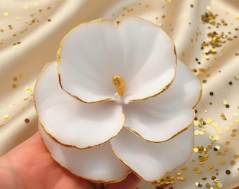 May include: A white flower-shaped candle with gold-painted edges on each petal and a central gold-painted stamen. The candle features layered, rounded petals that create a full, blooming appearance, with a smooth, matte finish on the white surfaces.