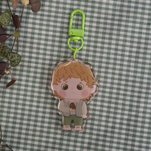 Op de afbeelding: Een heldere acryl sleutelhanger met een stripfiguur met lichtbruin haar, groene ogen en een groene broek. De sleutelhanger heeft een groene sluiting en een zilveren ring. Het personage houdt een klein bruin voorwerp vast.